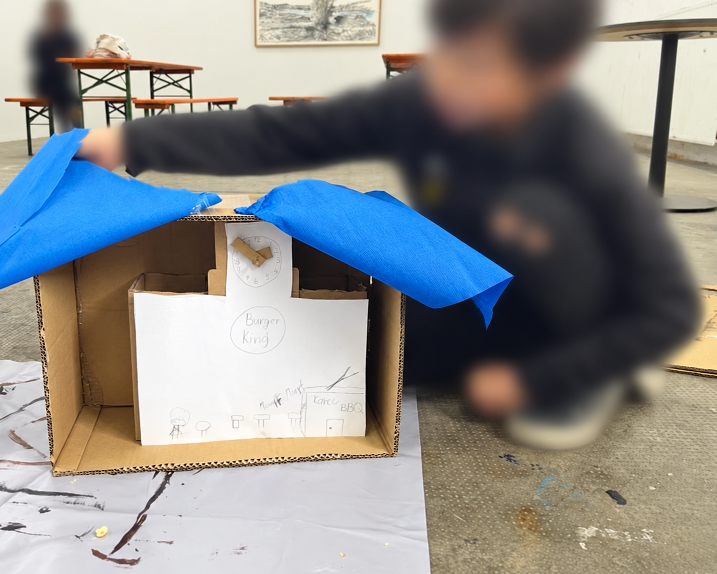 A child lifting the blue paper roof off a cardboard box theatre with a drawing of a Burger King restaurant and a cardboard clock
