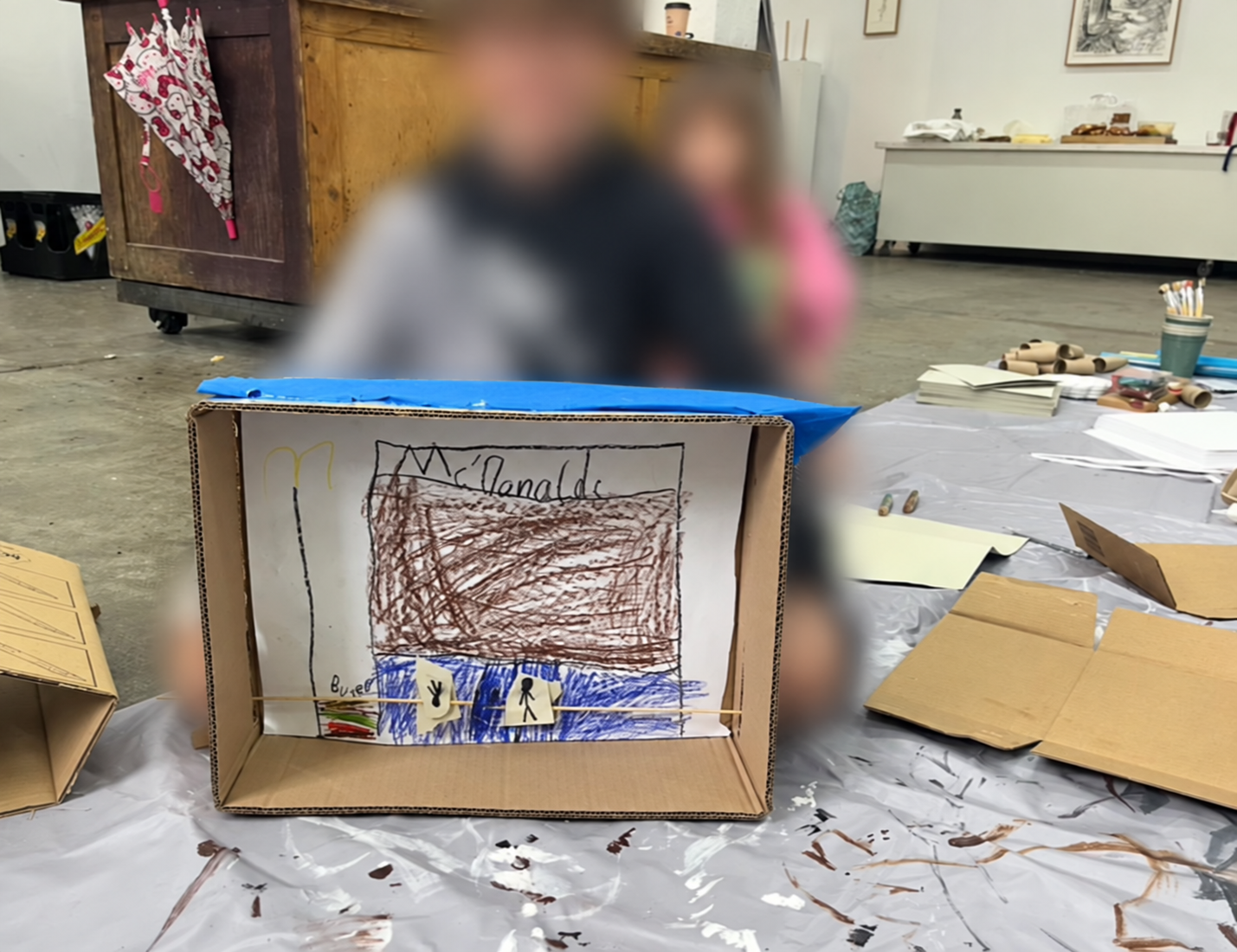 A cardboard box paper theatre with a child’s drawing of a McDonald’s restaurant and small paper figures on skewers