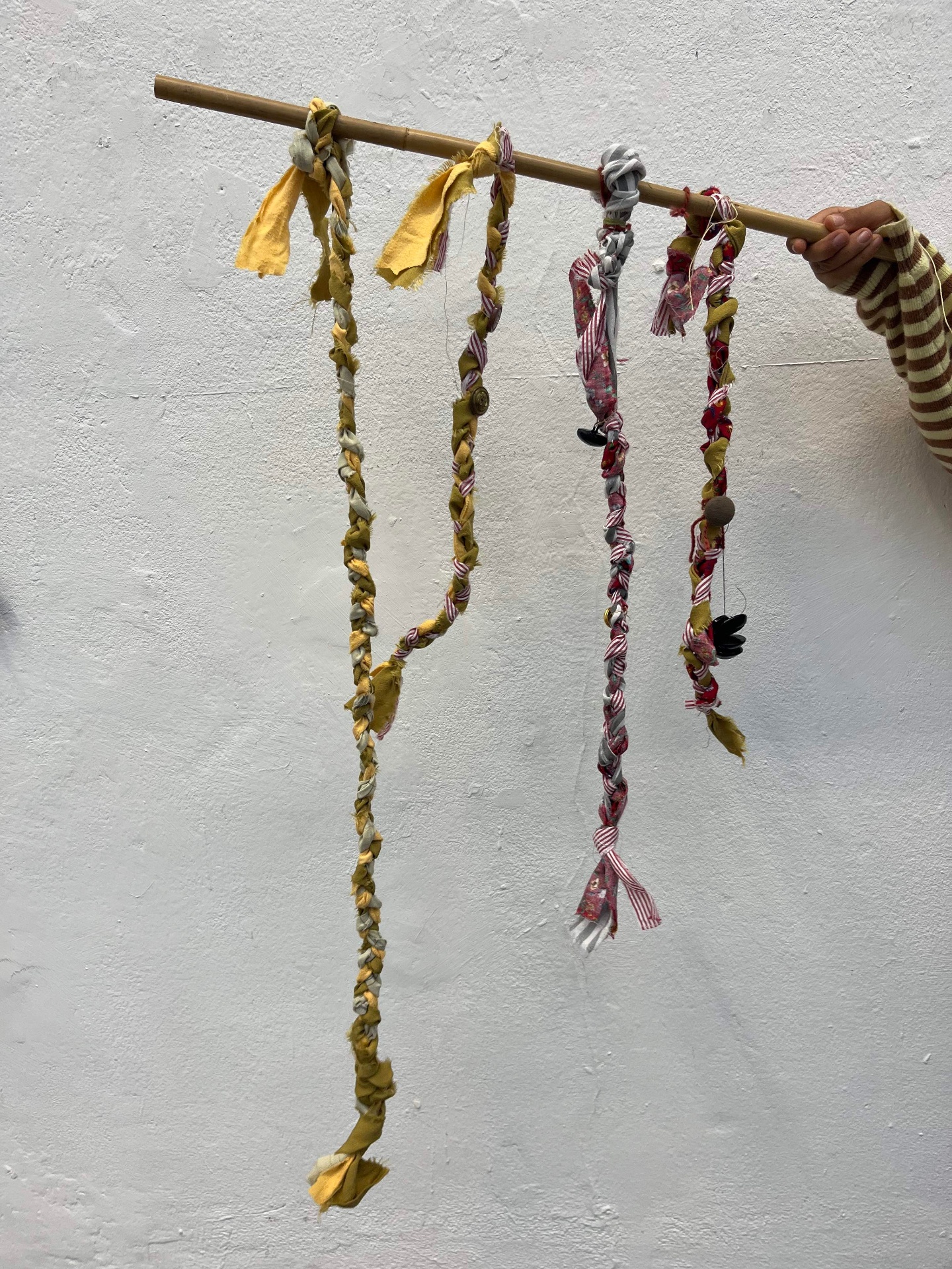 Braided fabric works in ochre, red and pink hanging from a bamboo pole held against a white wall